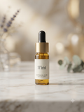 A small amber glass dropper bottle labeled 'Tint' with a white label and gold collar, standing on a light marble surface with a softly blurred warm-toned background.